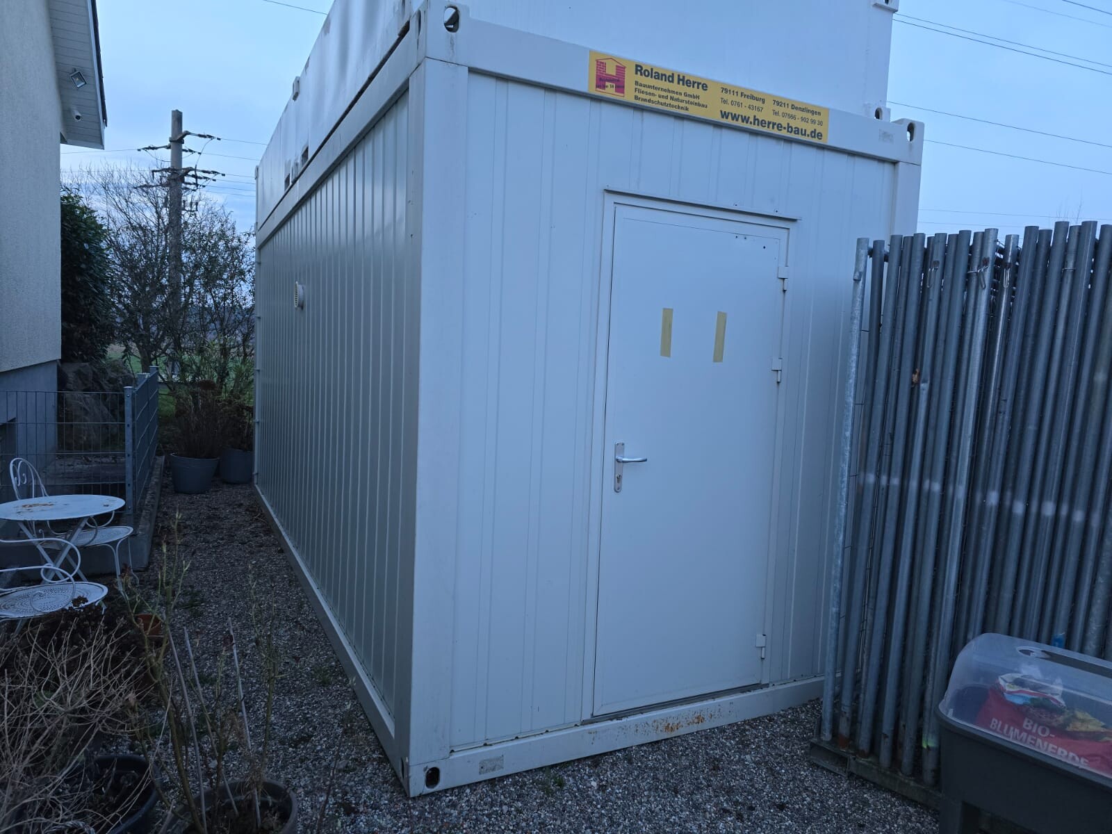 Weißer Baucontainer auf Kiesgrundstück neben Holzzaun; Gartenmöbel im Vordergrund.