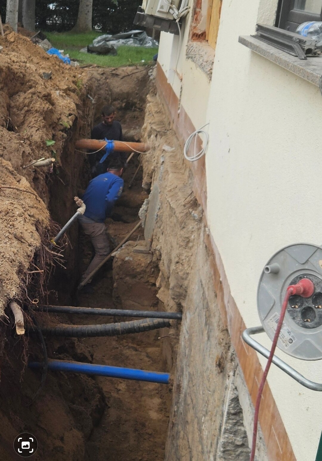 Zwei Arbeiter arbeiten in einem tiefen Baugraben neben einem Haus; sichtbare Rohre und Kabel.