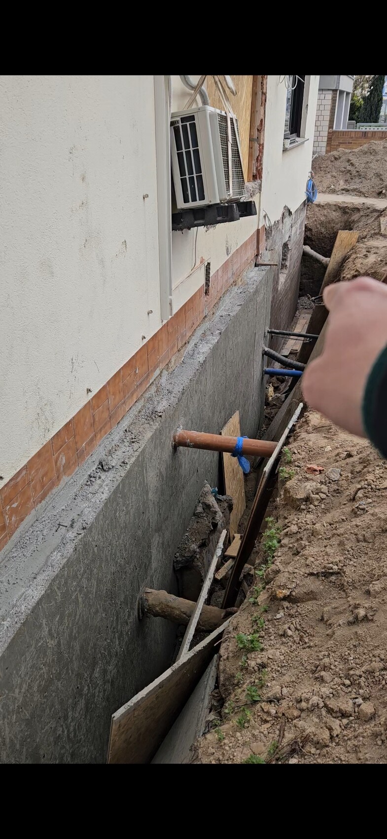 Baugrube neben Hausfassade mit freiliegenden Rohren, Bretter und einer Hand im Bild.