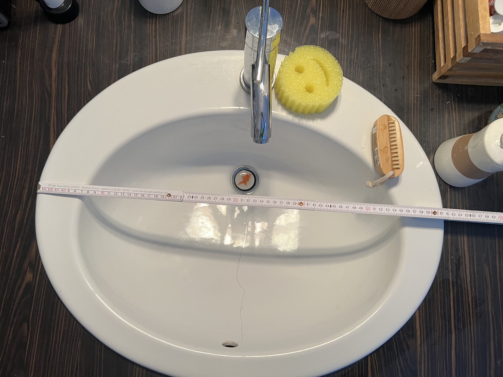 Weißes Waschbecken mit Maßband über der Breite, gelber Schwamm und Reinigungsbürste.