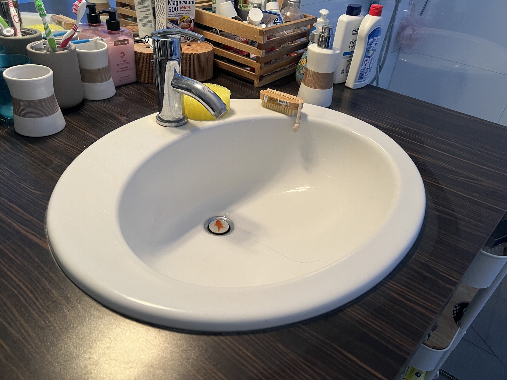 Weißes Becken mit Chromarmatur auf dunklem Holz, gelber Schwamm, Zahnbürsten im Hintergrund