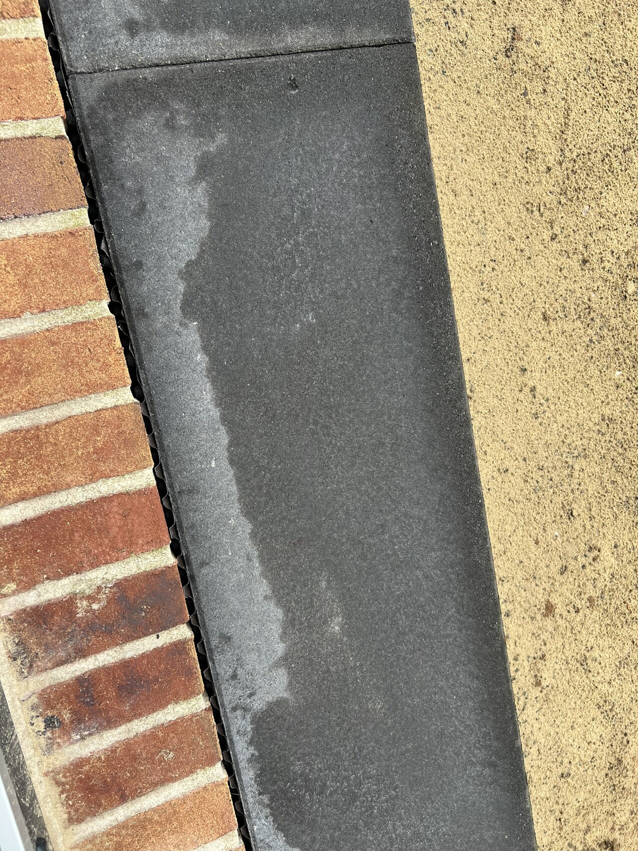 Nahaufnahme einer Dachkante: links Backstein, mittig dunkles Dachmaterial, rechts sandfarbiger Belag.