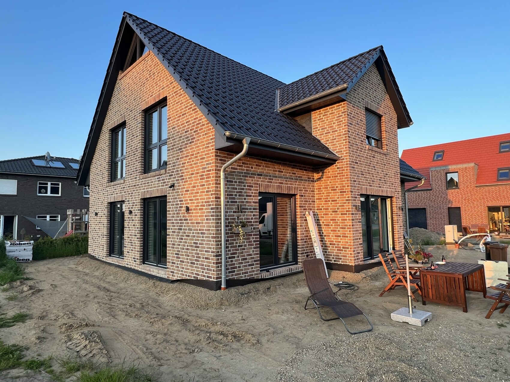 Zweistöckiges rotes Backsteinhaus im Bau mit dunklem Dach; Garten mit Holzmöbeln und Lounge-Sessel