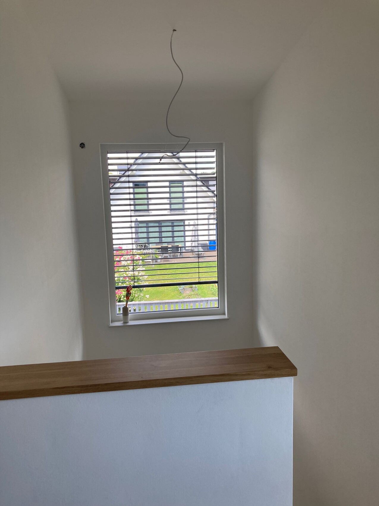 Heller Innenraum mit Fenster, Jalousie, Vase mit Blume auf Fensterbank, Holzhandlauf und lose Kabel.