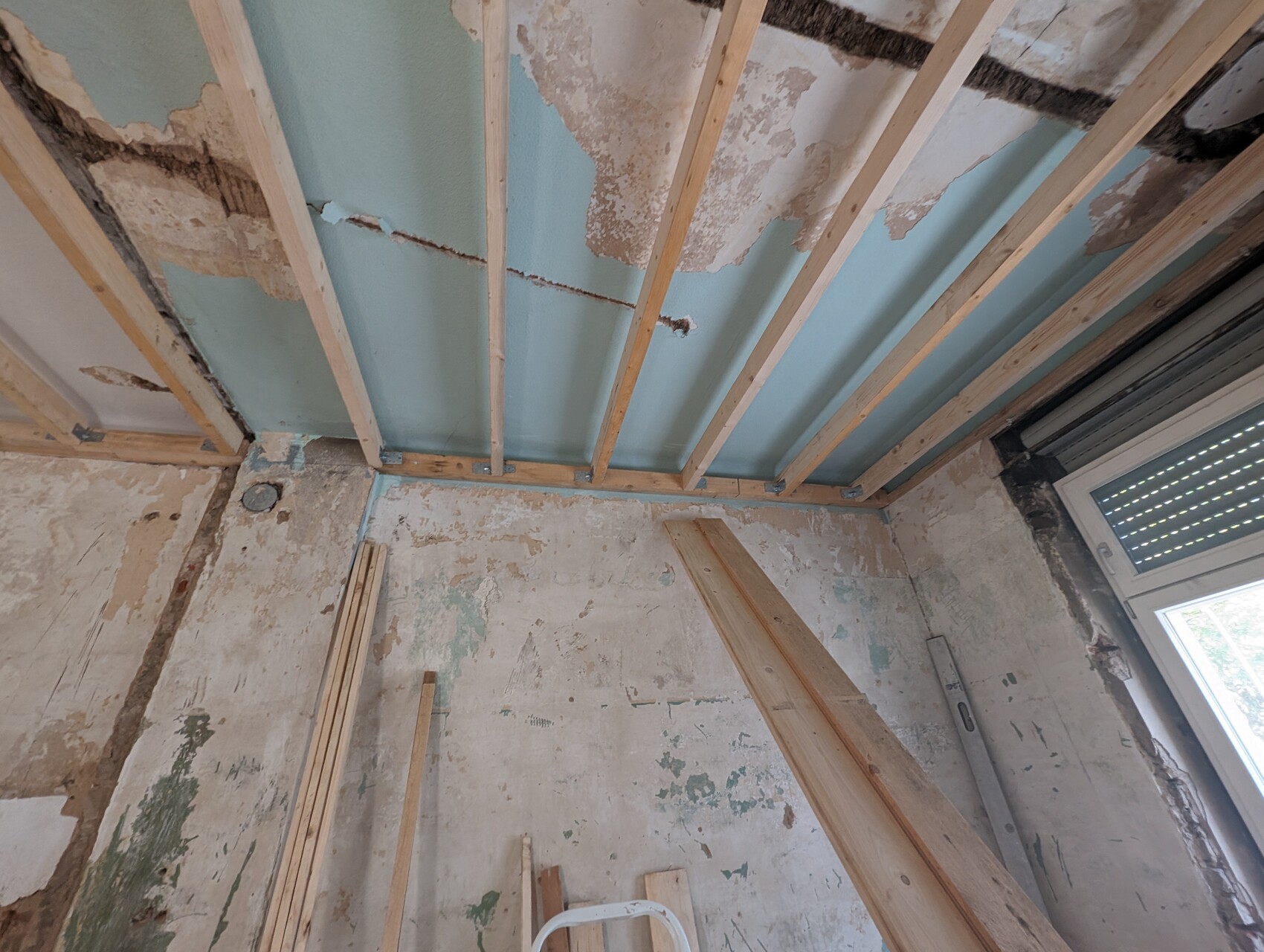 Raum im Renovierungszustand: Decke freigelegt mit Holzbalken, beschädigte Wände, Fenster rechts.