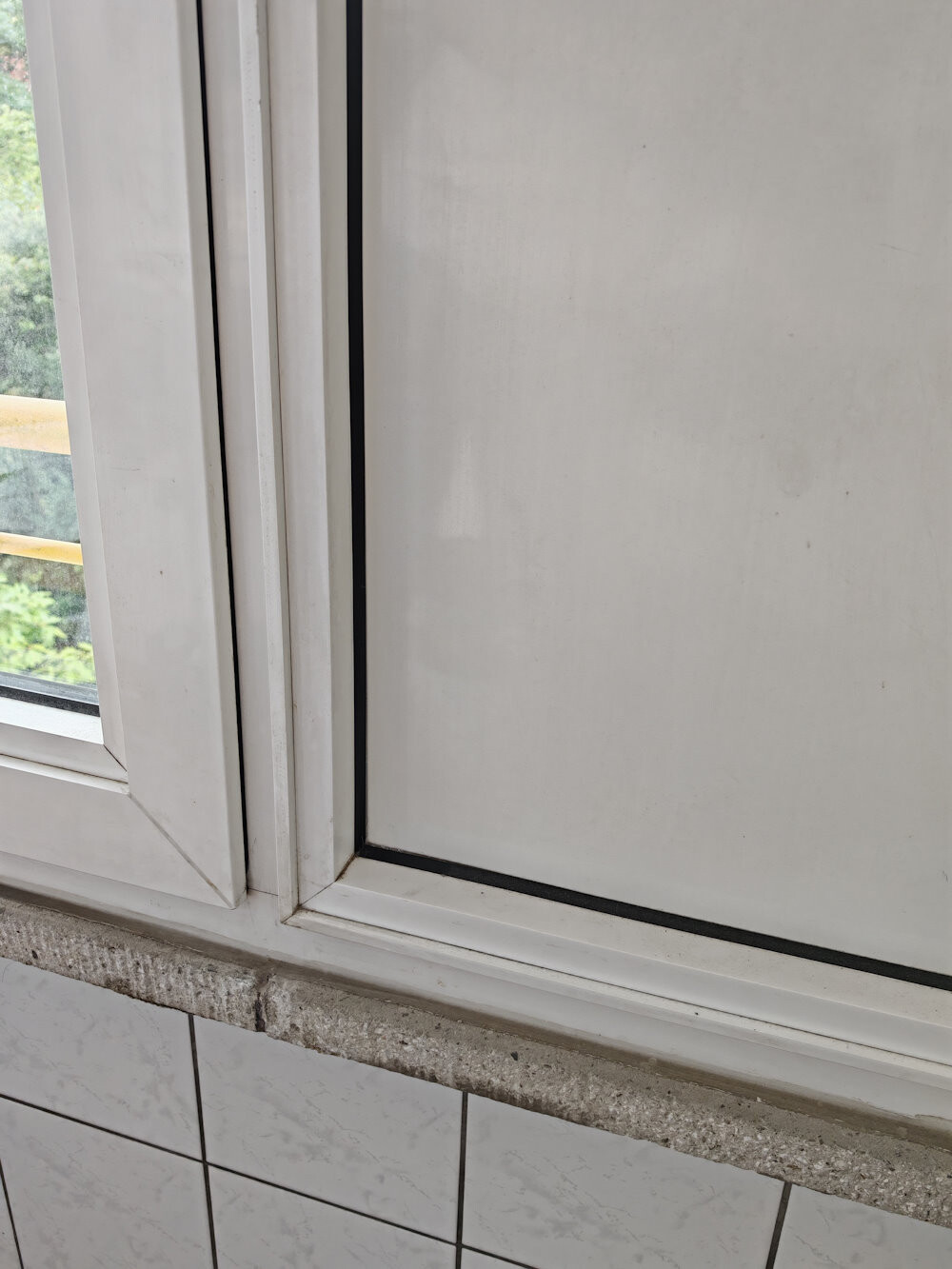 Weißer Fensterrahmen mit Glasscheibe, darunter Steinfenstersims und weiße Fliesen; außen grün sichtbar.