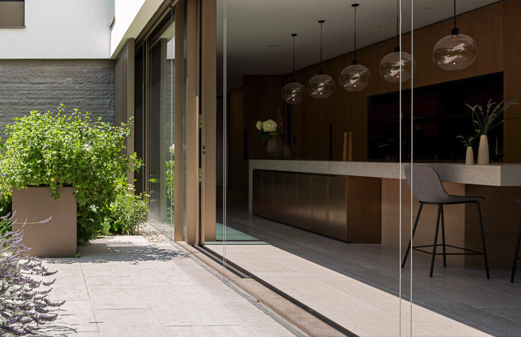 Moderne Küche mit Steininsel hinter Glas-Schiebetüren; außen grüne Pflanzen.