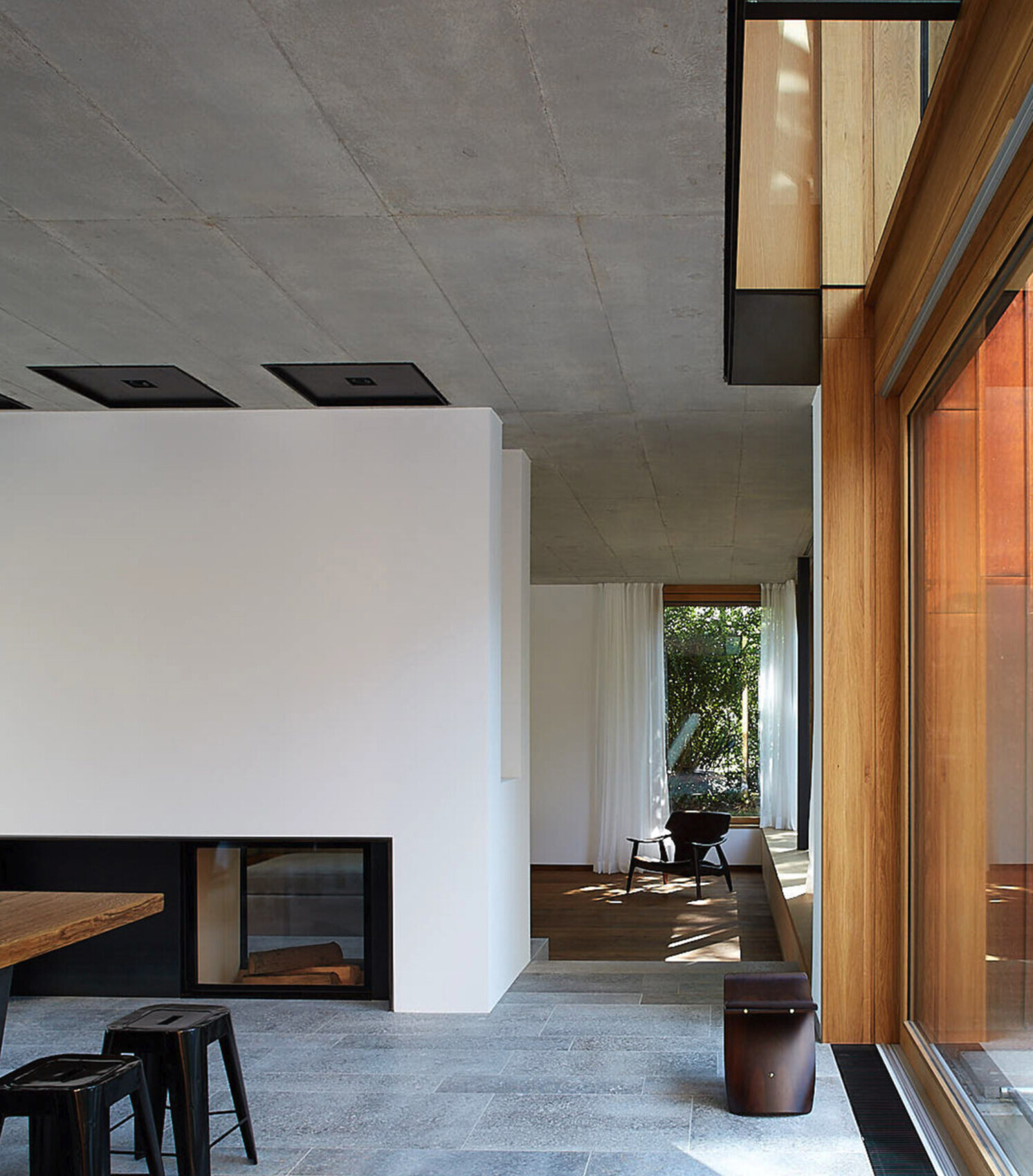 Moderner Innenraum mit grauer Beton-Decke, weißen Wänden, Holzrahmenfenstern, Kamin und Sitzbereich.