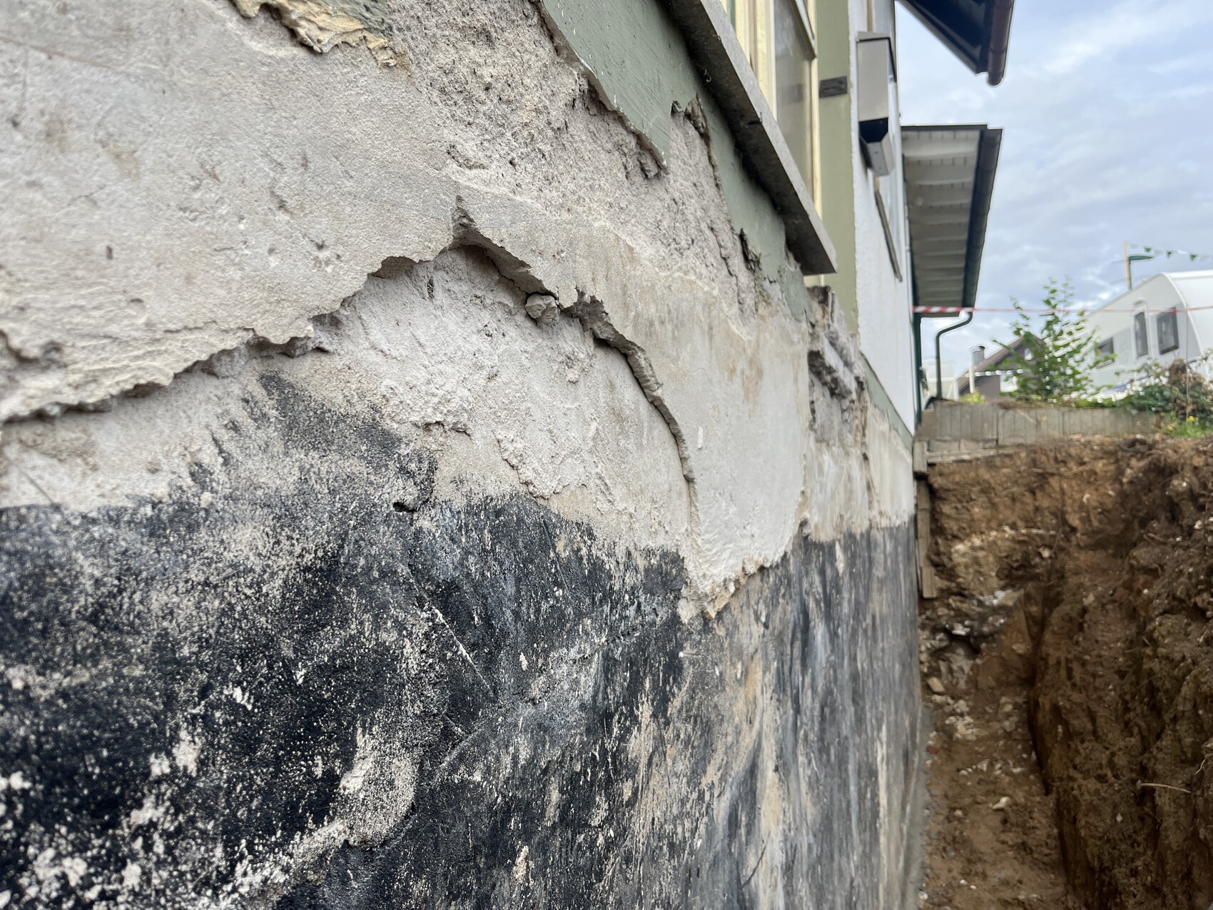 Außenwand eines Hauses mit abblätterndem Putz und freigelegtem Fundament neben einer Baugrube.