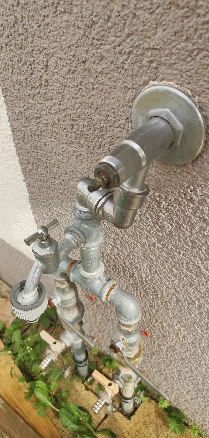 Außen-Wasserhahn mit mehreren Ventilen und Rohrleitungen an rauer Wand, Gartenboden.