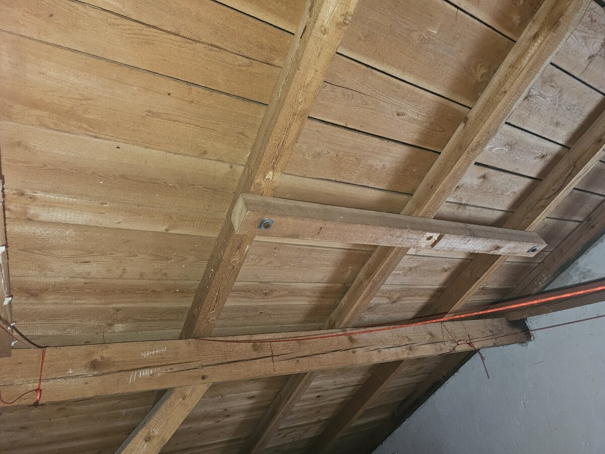 Unterseite eines Holz-Dachstuhls mit Balken, Holzbrettern und sichtbaren Kabeln.