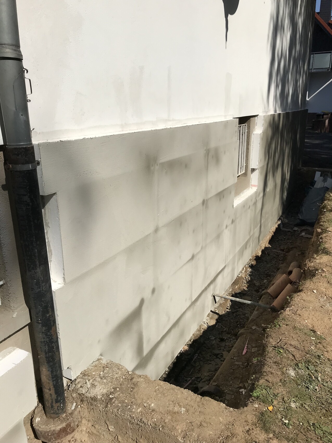 Außenwand eines Gebäudes mit grauer Fundamentverkleidung, Baugrube daneben und links Abflussrohr.