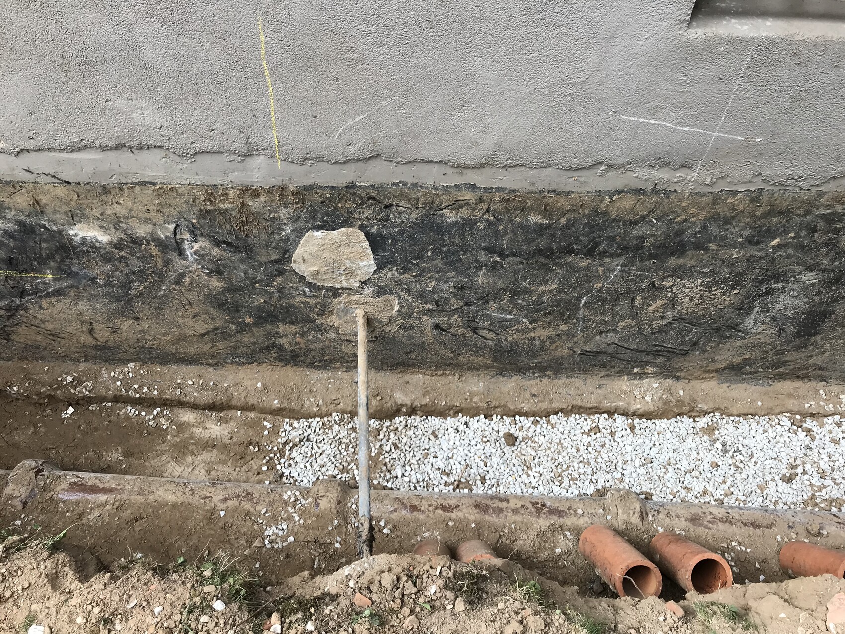 Aushub neben einer Betonwand: Kiesbett, orange Tonrohre, Stabpfahl und Betonreste.