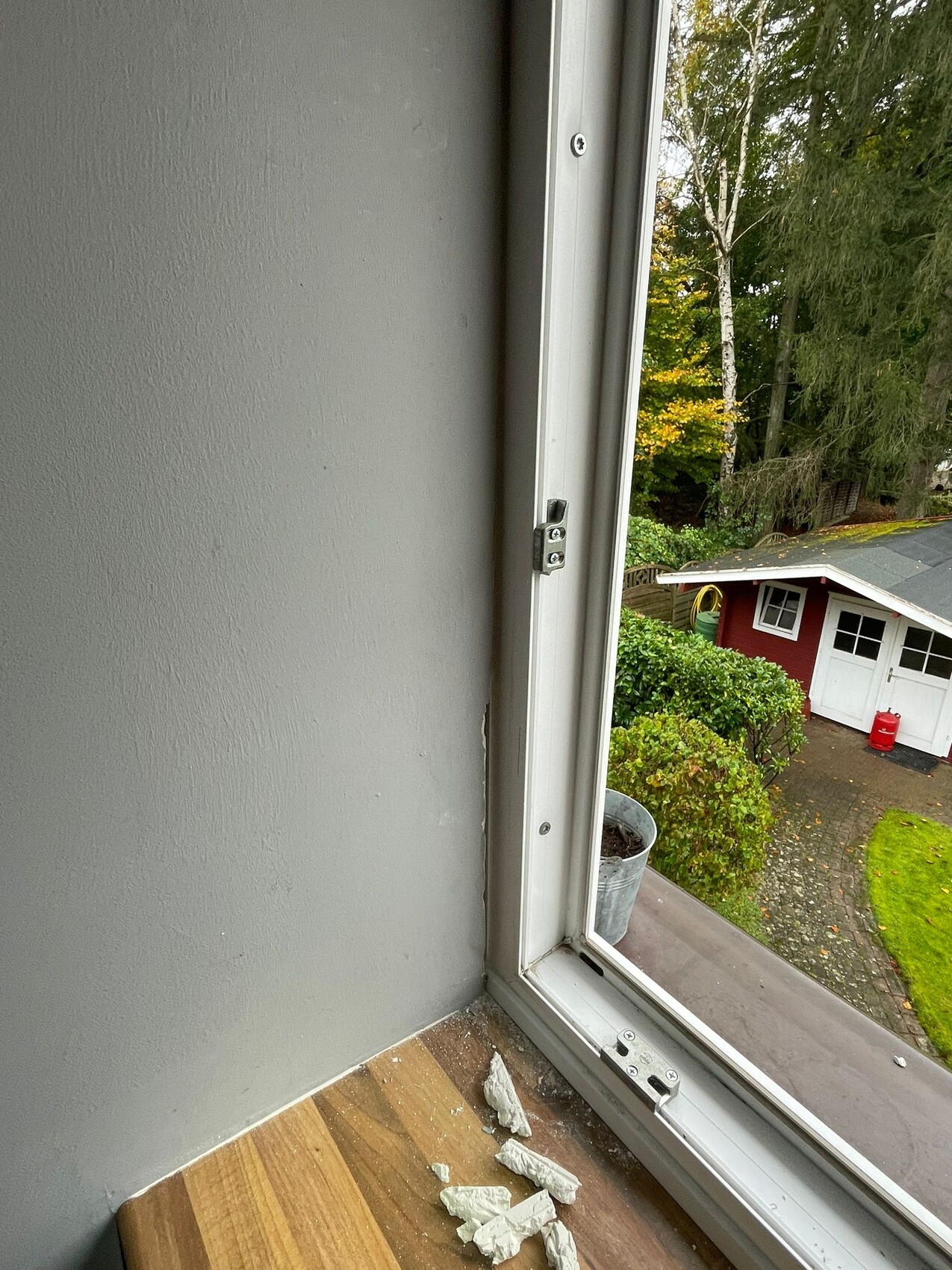 Fensterblick: Innenraum mit grauer Wand, Holzboden und Gipsstücke; außen Garten mit roter Hütte.