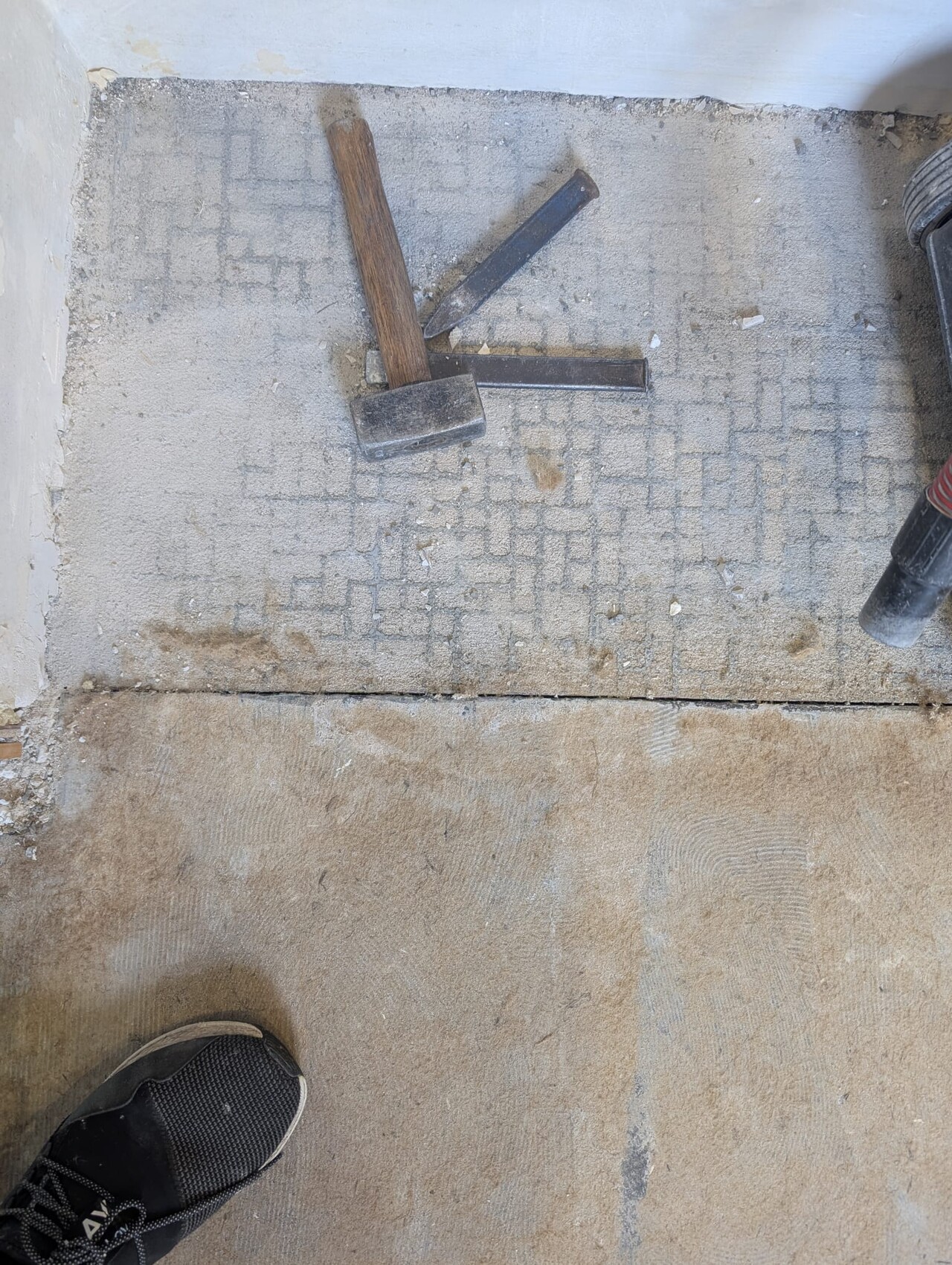 Hammer und Meißel liegen auf staubigem Betonboden einer Bauzone; Schuh am Rand sichtbar