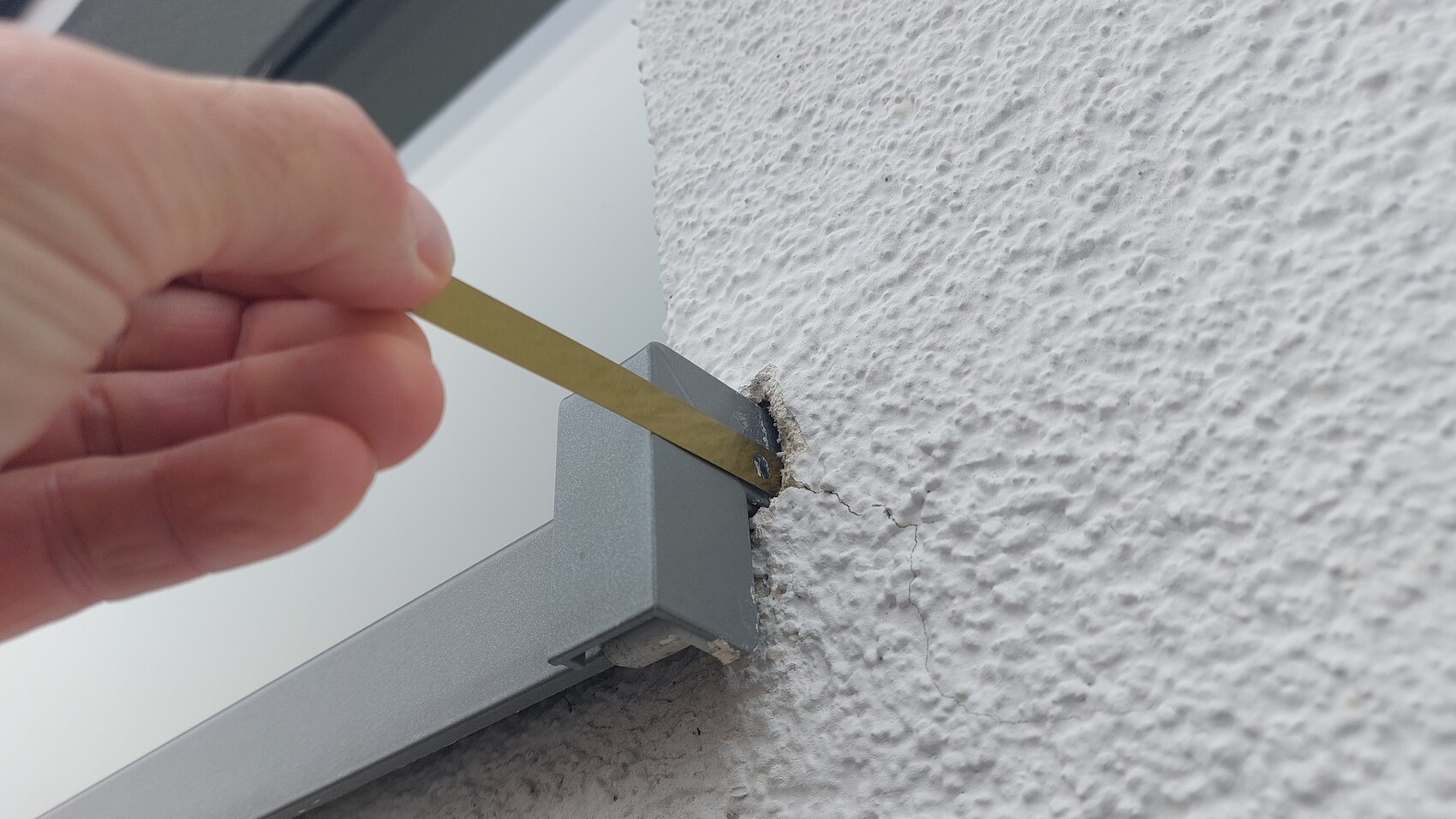 Hand hält einen Metallkeil zwischen grauem Fensterrahmen und rauer weißer Wand; Riss sichtbar.