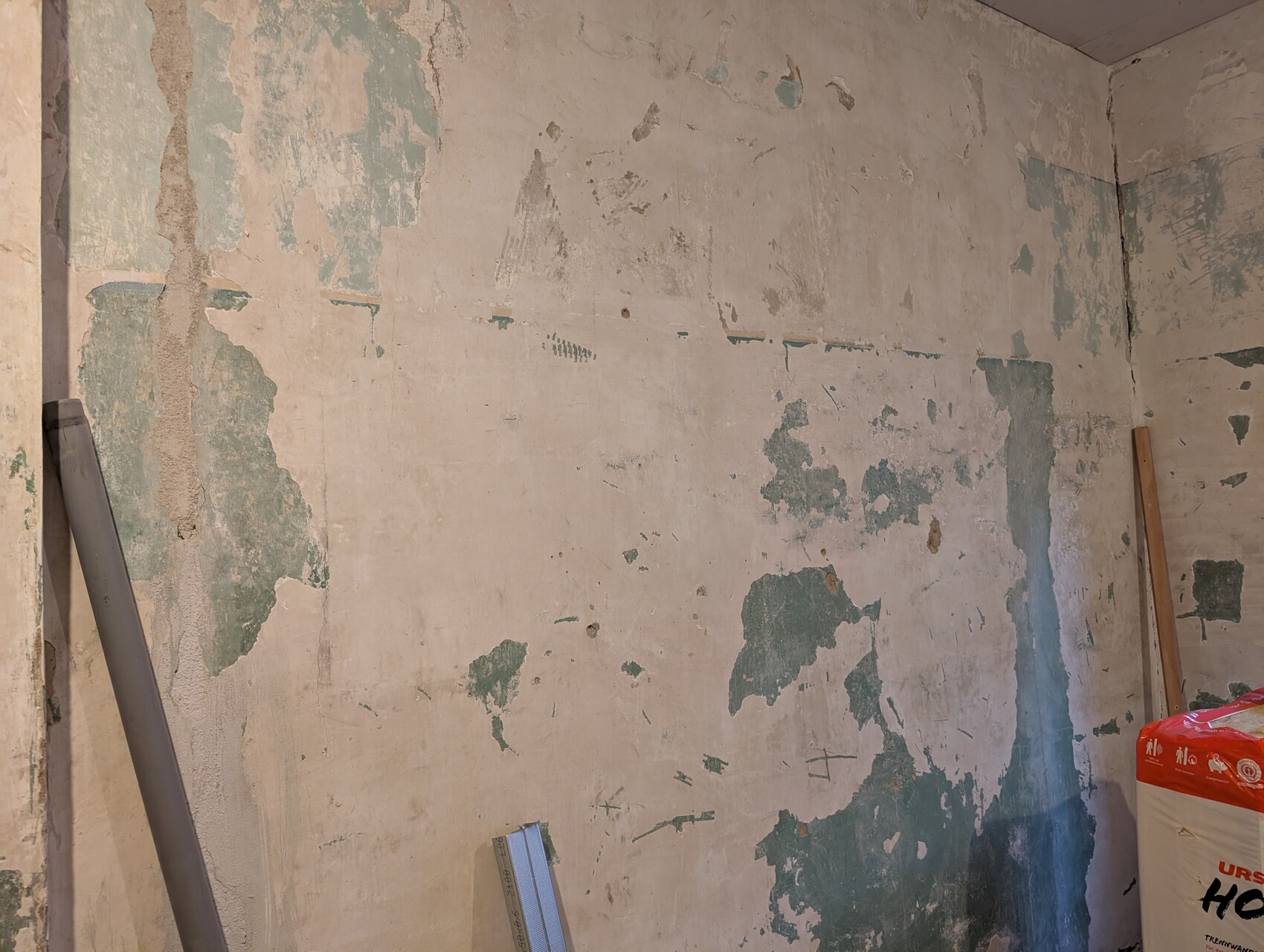 Verputzte Wand mit abblätternder grün-blauer Farbe; linkes Rohr, Bauutensilien und Baubeutel rechts unten.