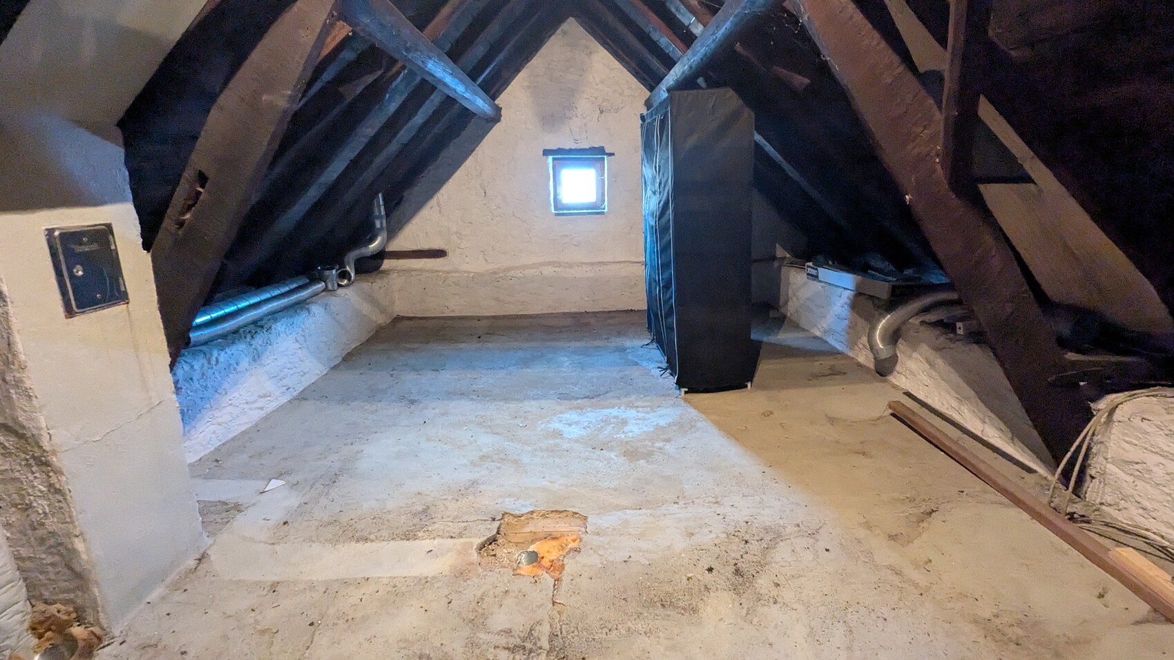 Dachboden mit Holzbalken, kleinem Fenster, schwarzem Schrank, Rohren und Bauholz.