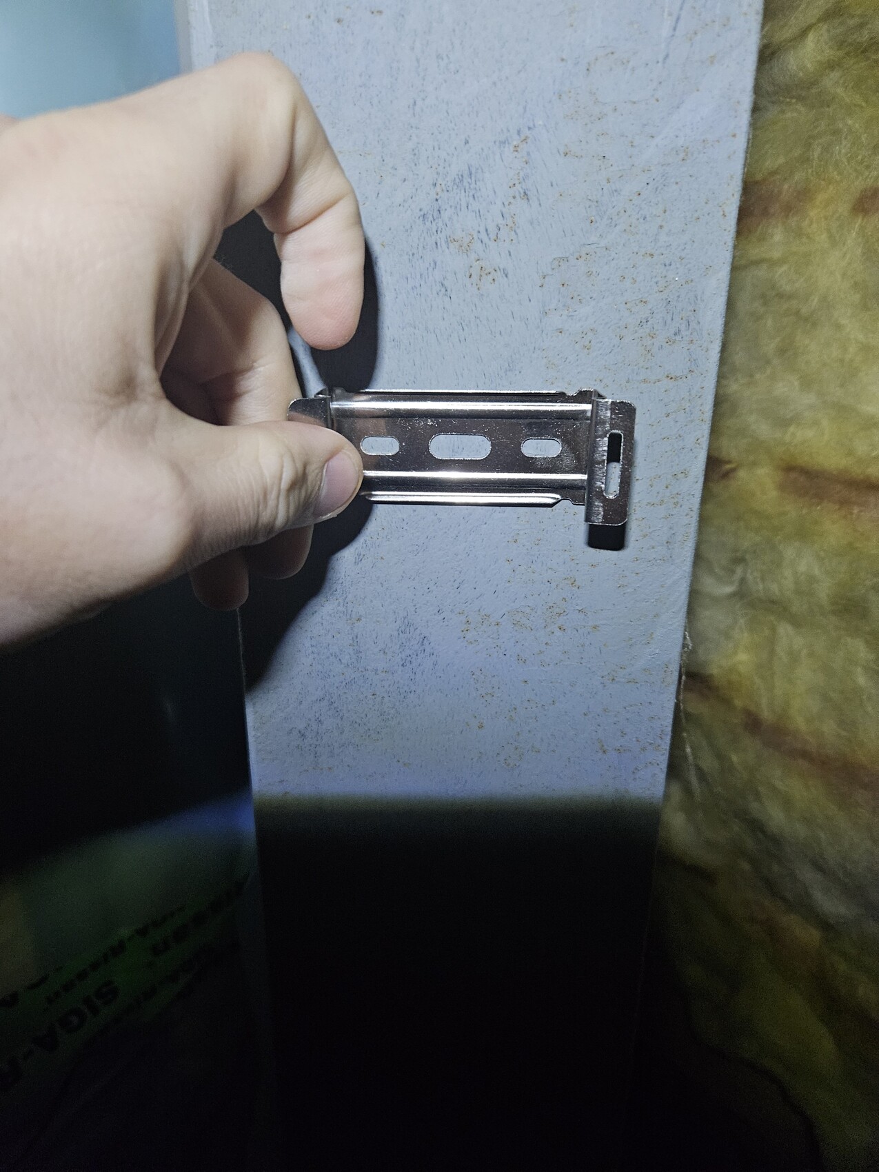 Hand hält eine flache Metallhalterung mit drei ovalen Löchern vor einer bläulich-grauen Metallfläche