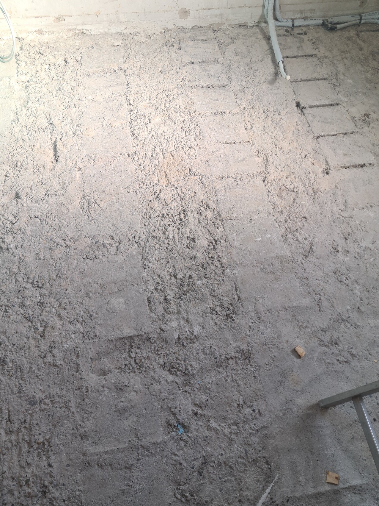 Baustellenboden mit Staub, Kies und Spuren, Rohre an der Wand sichtbar.