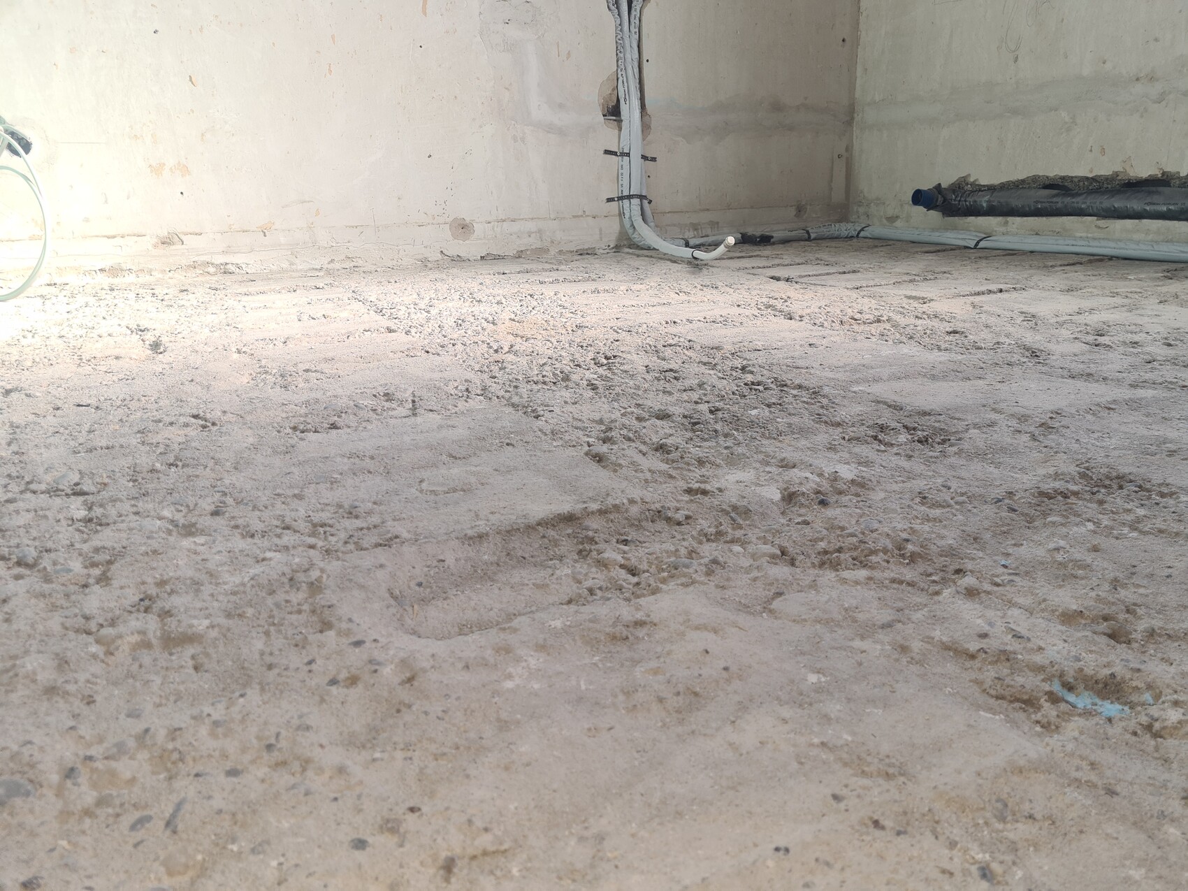 Rauer Betonboden einer Baustelle, Staub auf dem Boden, Rohre und Kabel an der Wand.