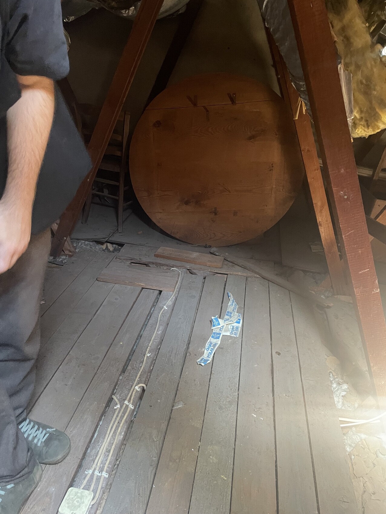 Verstaubter Dachboden, zentral ein großer runder Holzkörper, Holzboden, links eine Person sichtbar.