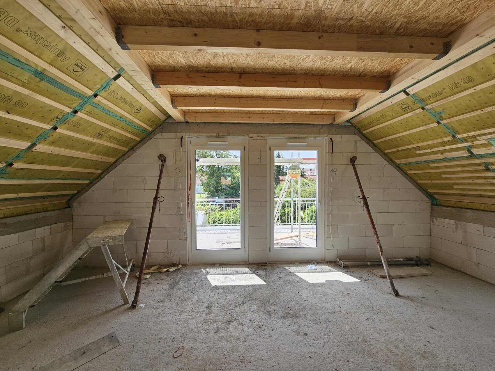 Dachgeschoss-Raum im Bau mit Holzbalken, Glas-Türen nach draußen und Stützpfosten.