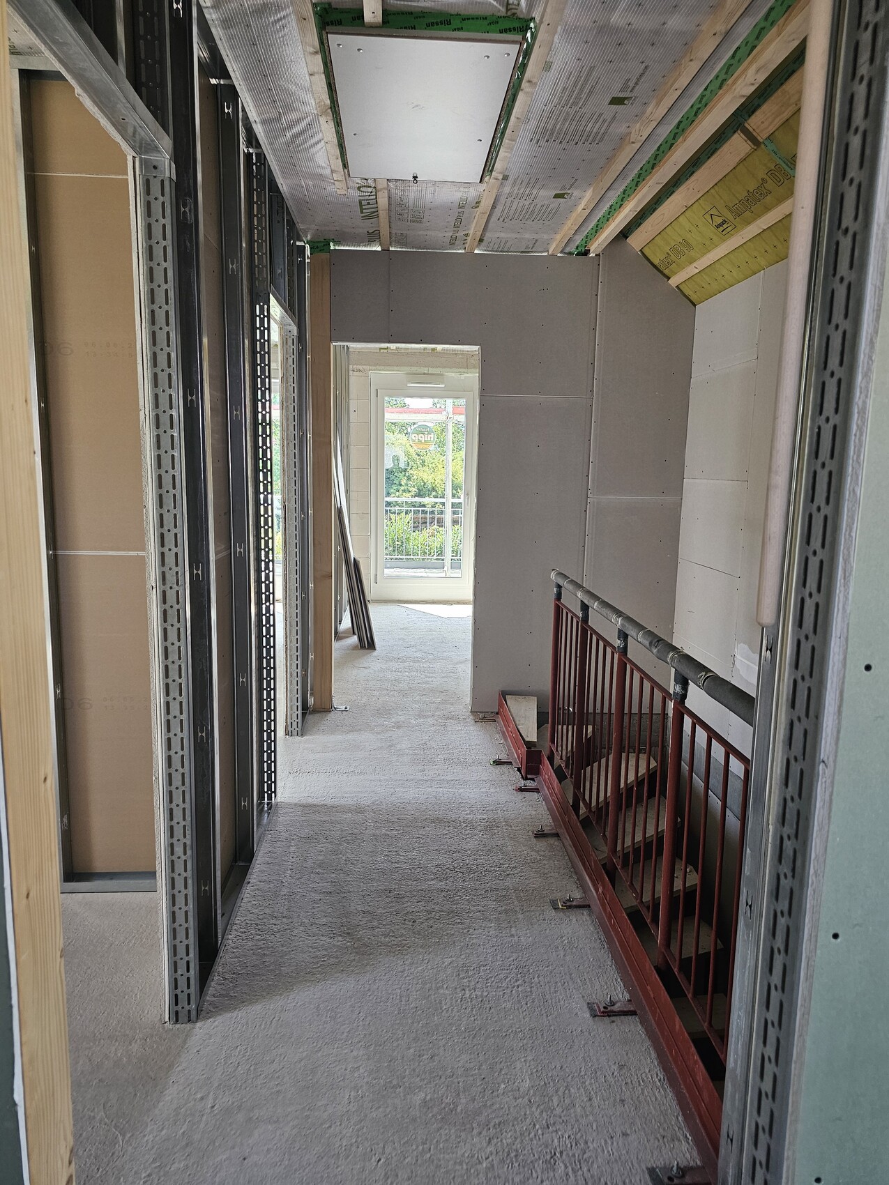 Rohbau-Flur: links Stahlständerwerk, rechts rotes Geländer, Endfenster mit Aussicht.