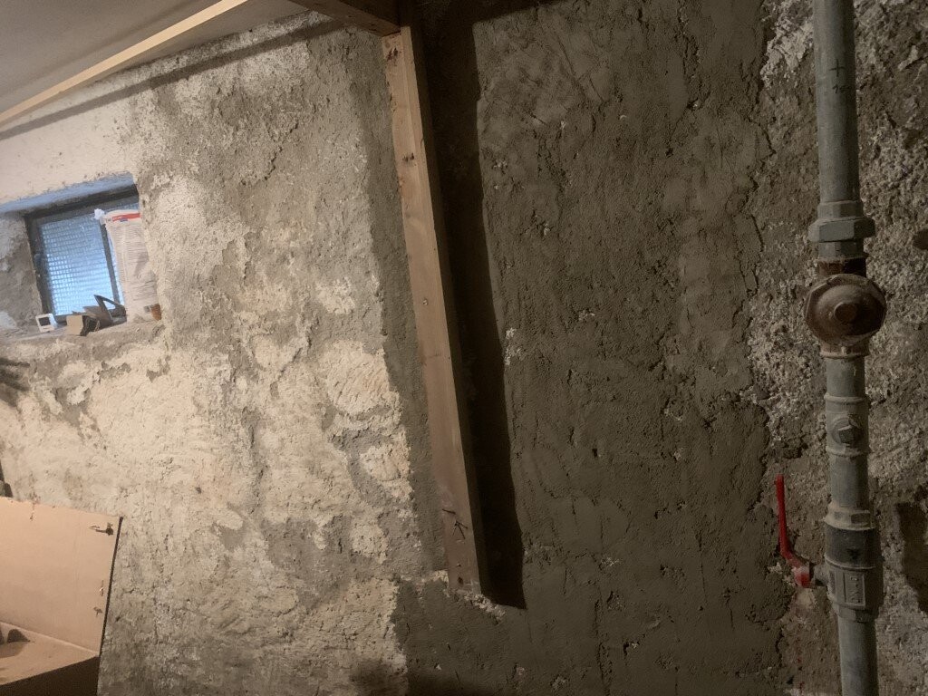 Rohbau-Keller: raue Betonwand, Holzstütze, Rohrleitung mit Ventil rechts, kleines Fenster.