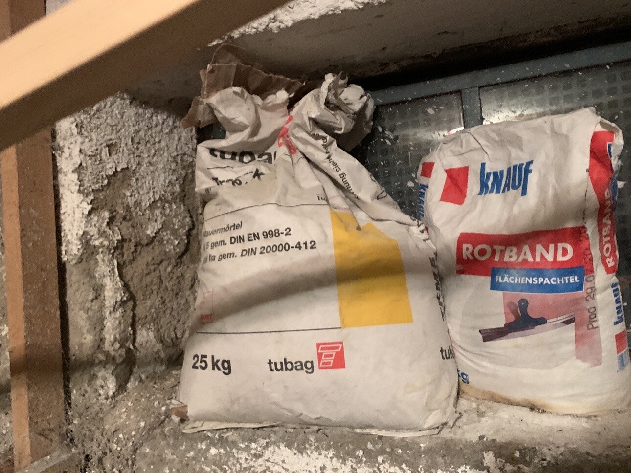 Zwei Bau-Säcke (25 kg) von tubag und Knauf Rotband stehen im Keller neben einer Wand.