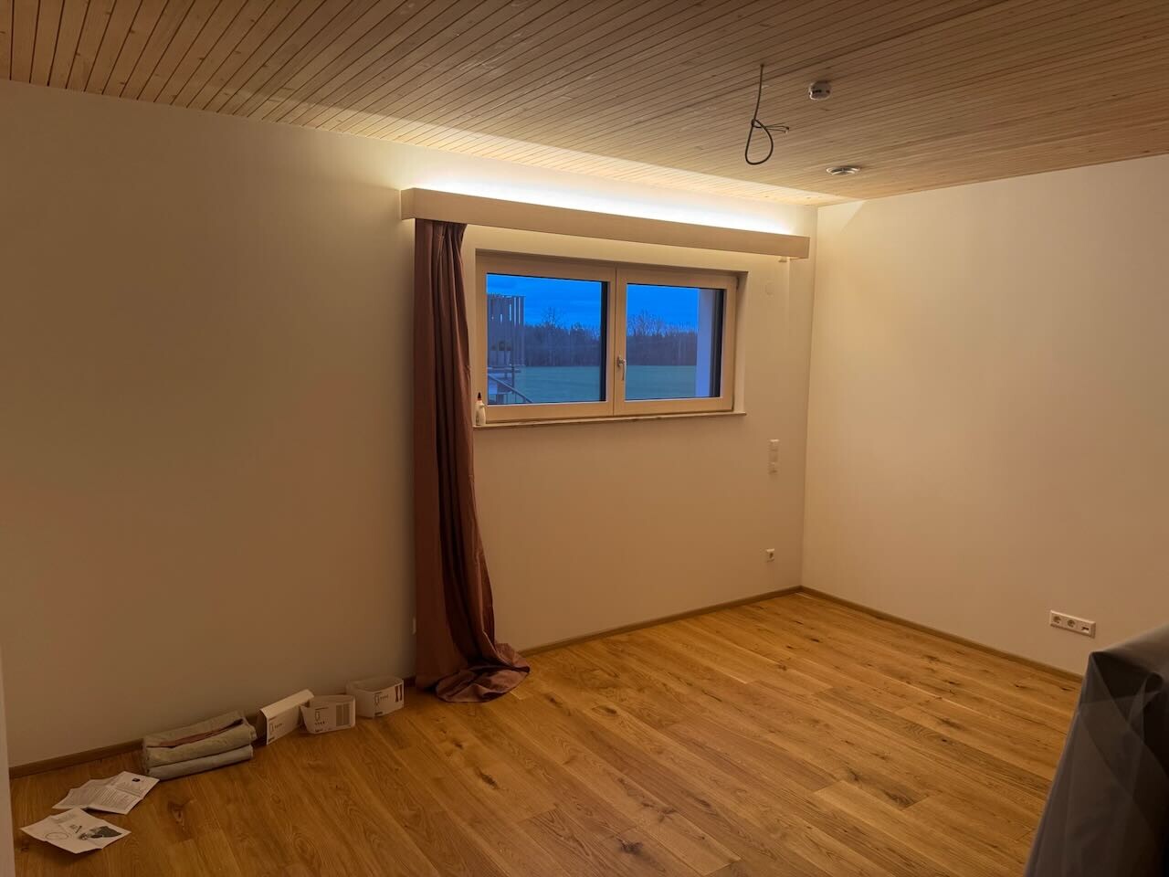 Leeres renoviertes Zimmer mit Holzfussboden, Fenster, Vorhang und Decke aus Holzleisten.