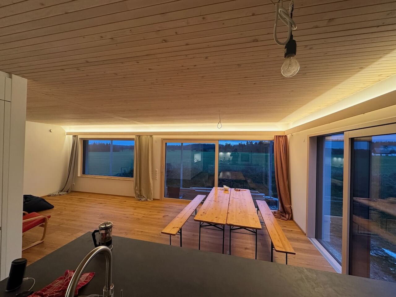 Helles Wohnzimmer mit Holzboden und Holzdecke, Esstisch mit Bänken, Glasfenster mit Blick aufs Feld.