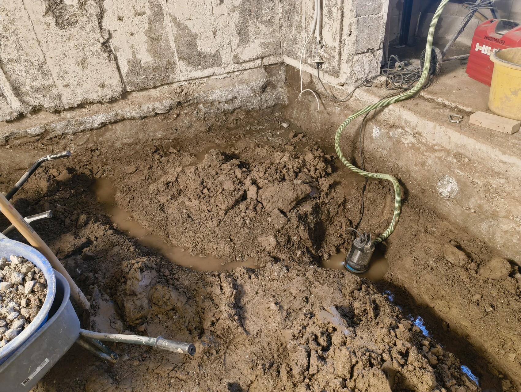 Ausgehobene Baugrube neben Fundamentwand; Pumpe mit Schlauch, Schubkarre voller Kies.