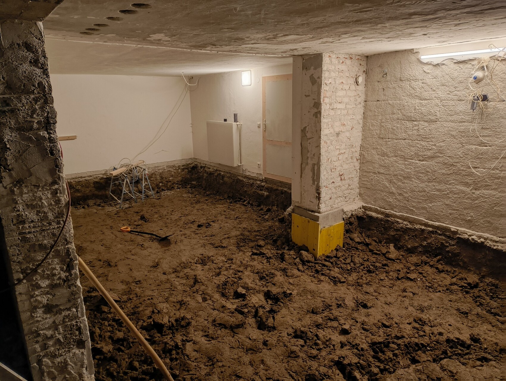Kellerbaustelle: Erdreichboden, gelber Stützenfuß, raues Mauerwerk, Provisoriumbeleuchtung.