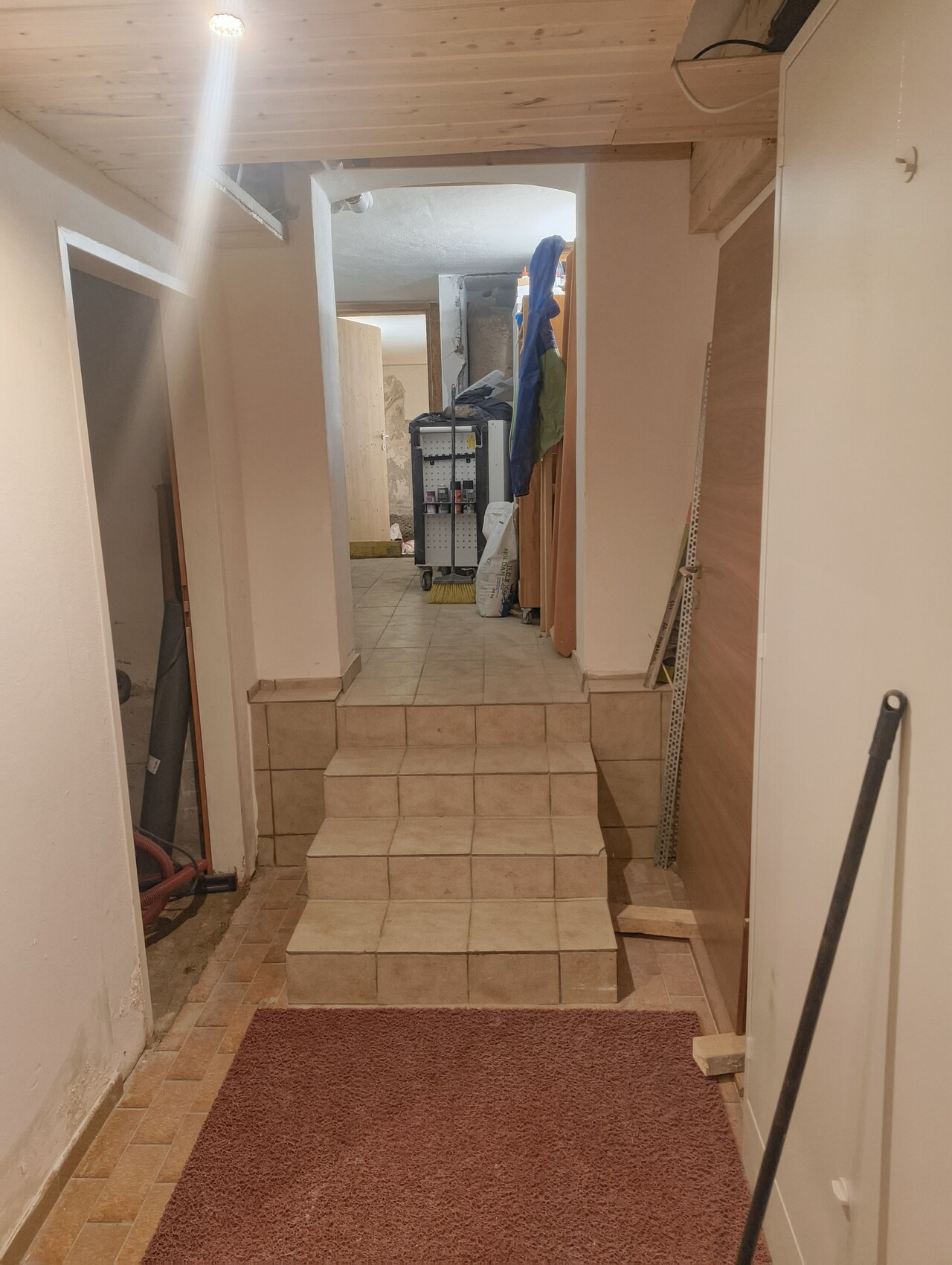 Heller Kellerflur mit dreistufigen Fliesenstufen, Reinigungswagen und Mantelgarderobe im Hintergrund.