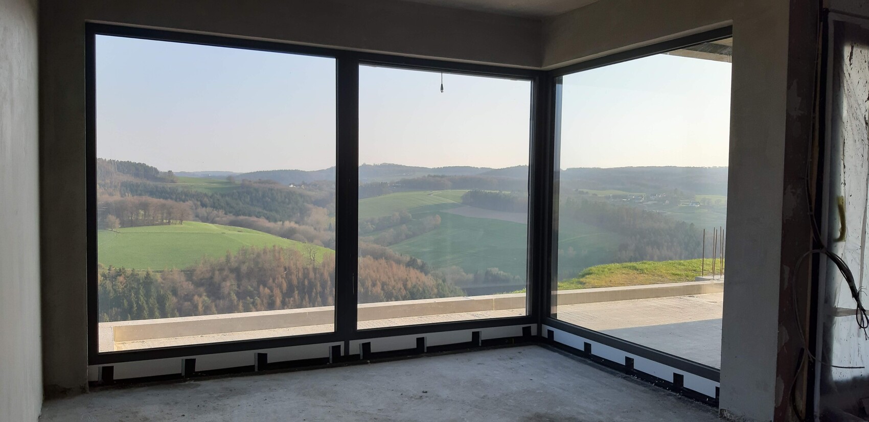 Unfertiger Innenraum mit großen Eckfenstern und Aussicht auf grüne Landschaft