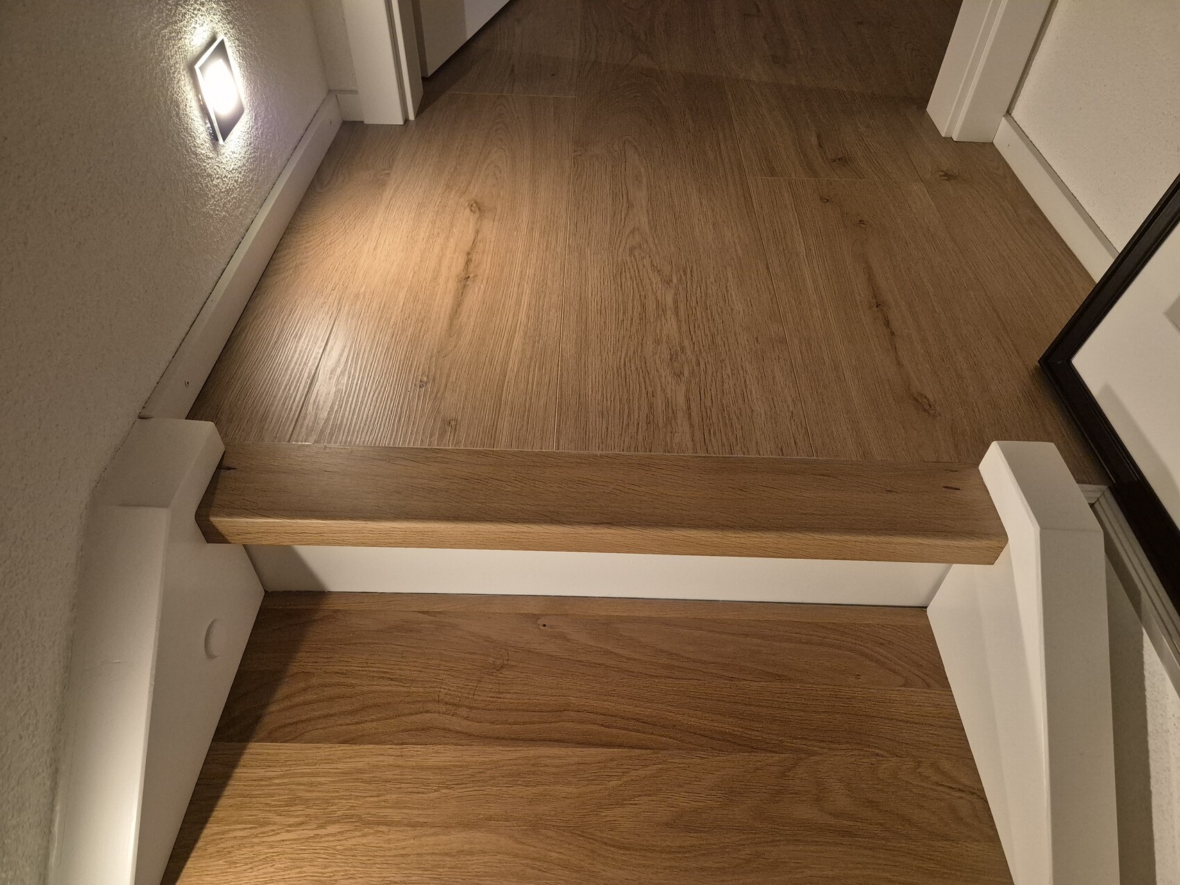 Blick auf eine moderne Holztreppe im Innenbereich mit Wandlicht und weißer Umrandung