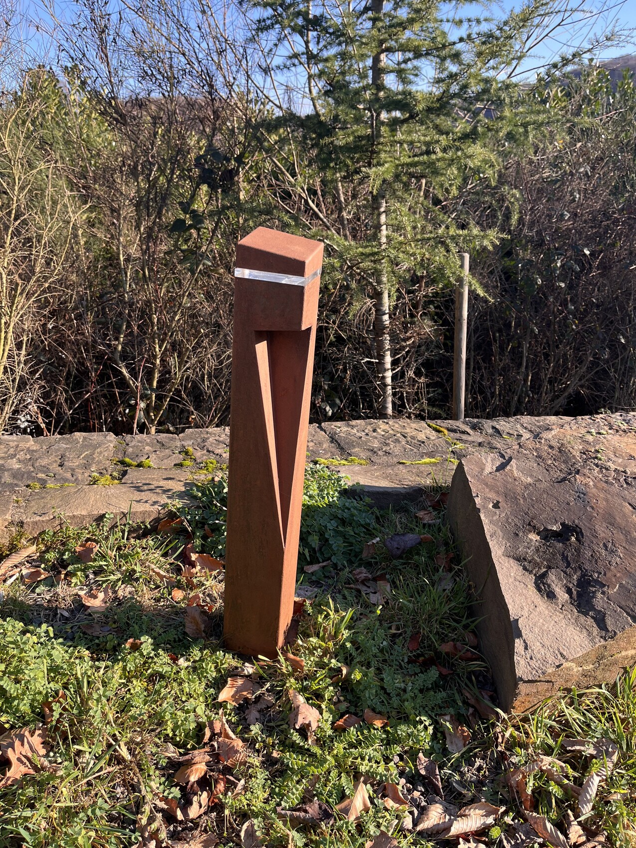 Rostiger Metallpfosten im Garten neben Steinmauer, von Pflanzen umgeben.