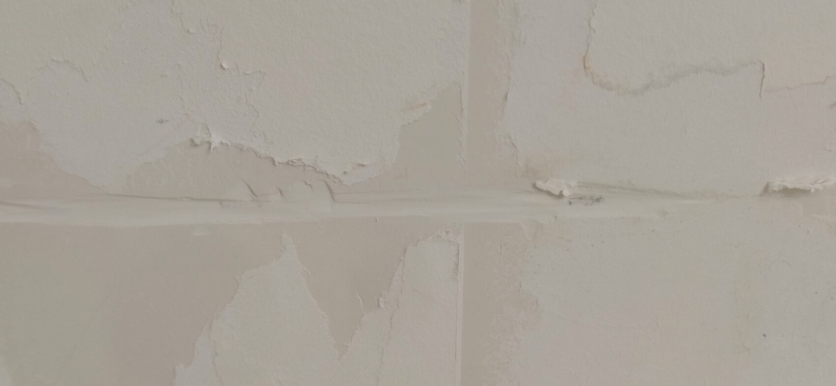 Weiße Wand mit abblätternder Farbe, Rissen und einer horizontalen Nahtstelle.