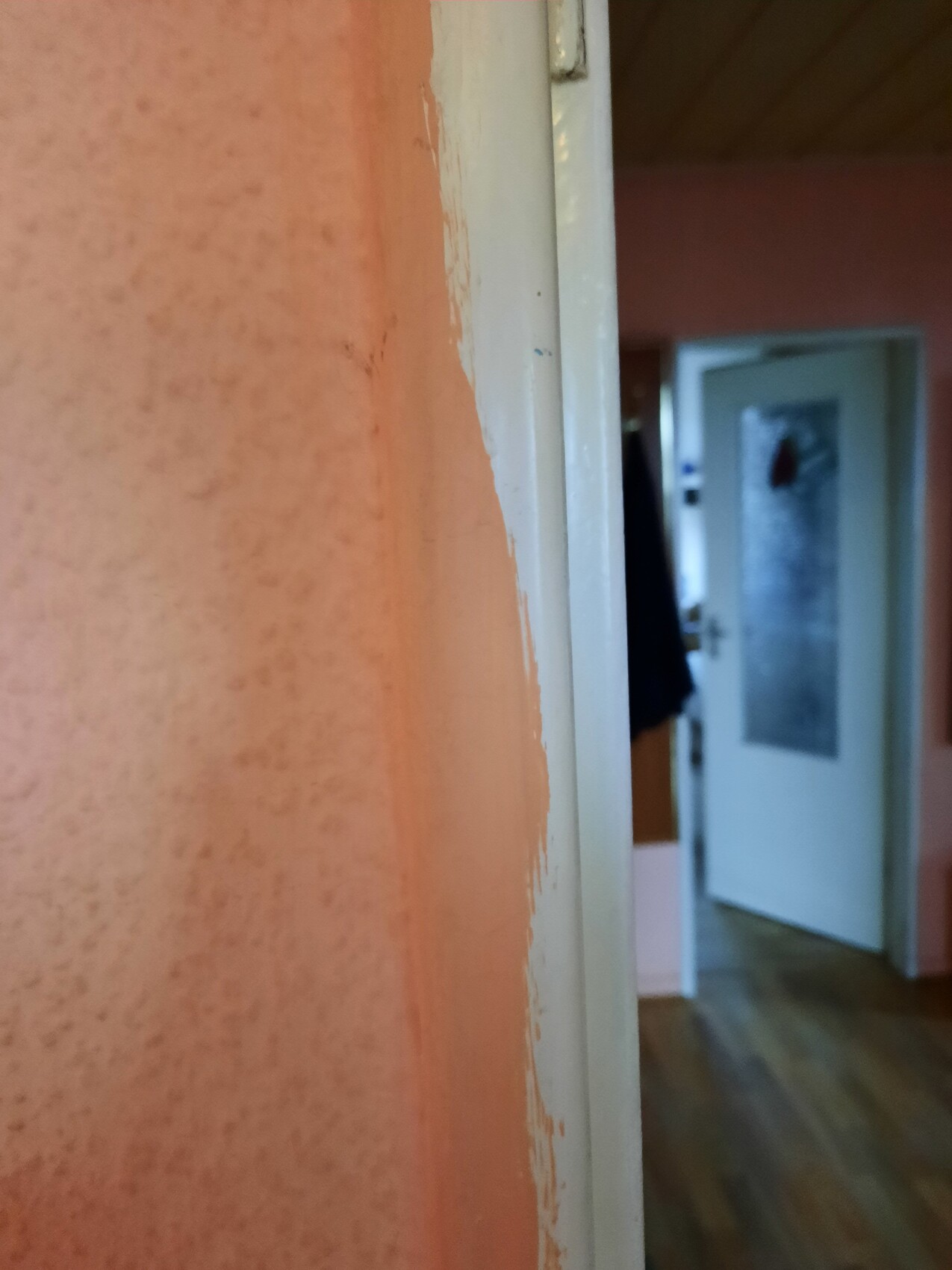Nahaufnahme einer orangefarbenen Wand mit abblätternder Farbe neben weißem Türrahmen; offene Tür im Hintergrund.