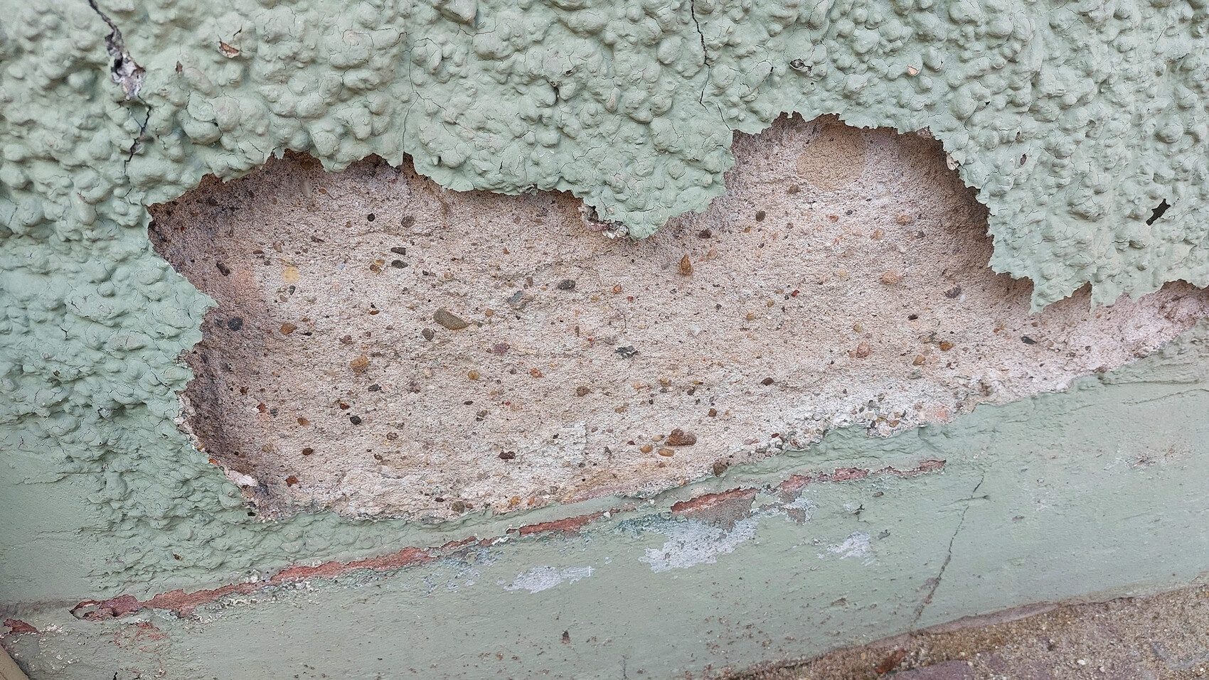 Nahaufnahme grün gestrichener Wand mit abgeplatzter Farbe und grobem Putz; Kiesel sichtbar