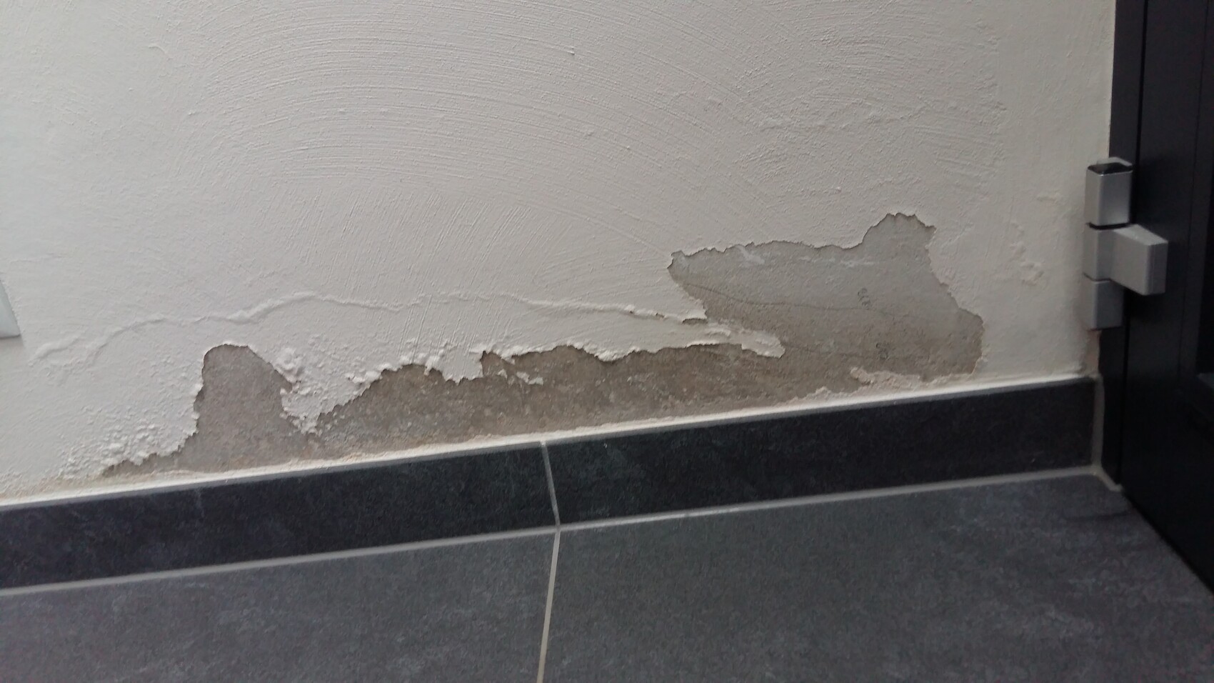 Wand mit abgeblättertem Putz und freiliegendem Untergrund über dunklen Fliesen; Türrahmen rechts.