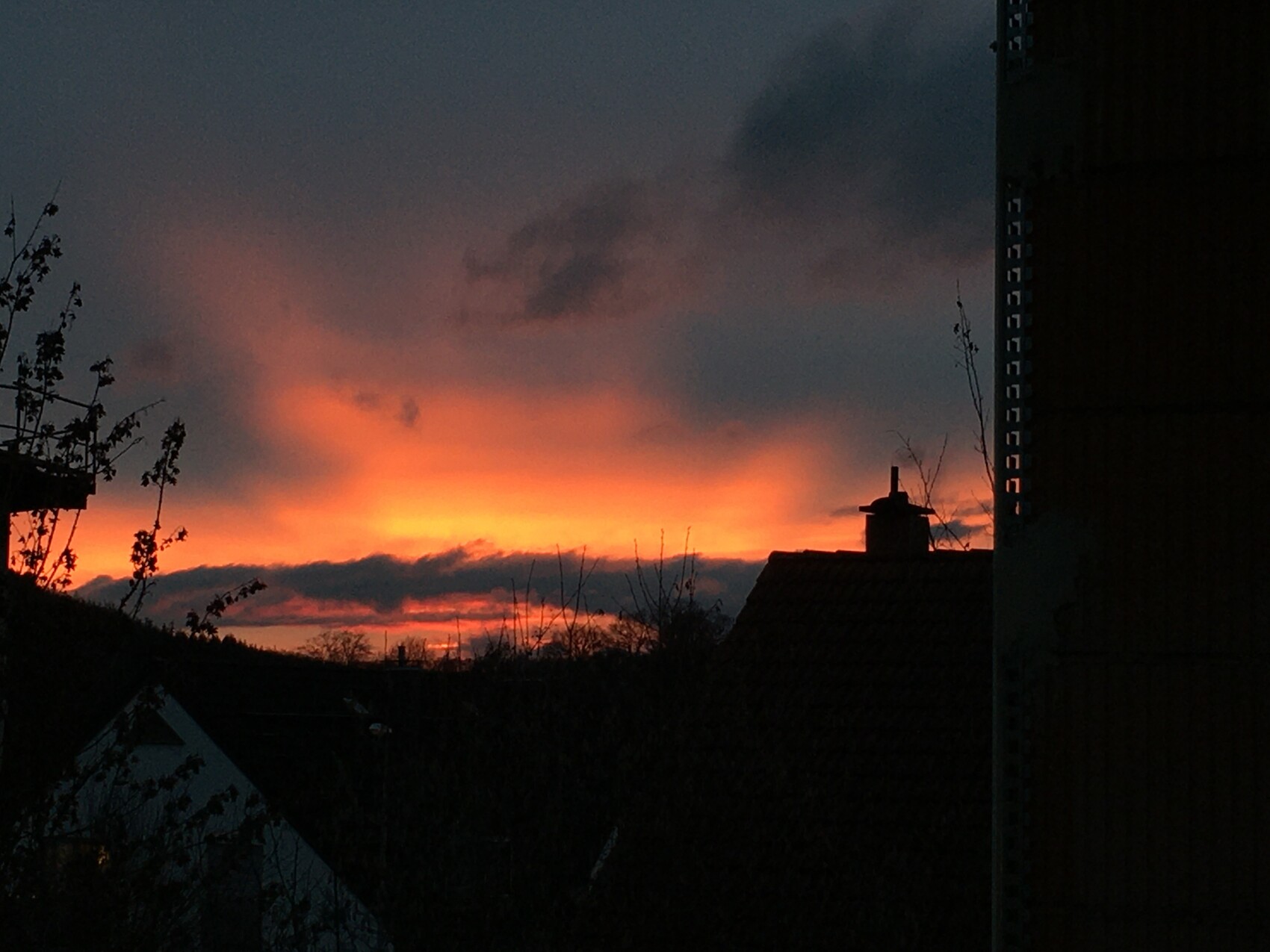 Sonnenuntergang hinter Hausdächer; orangefarbener Himmel, dunkle Gebäudesilhouetten und Bäume.