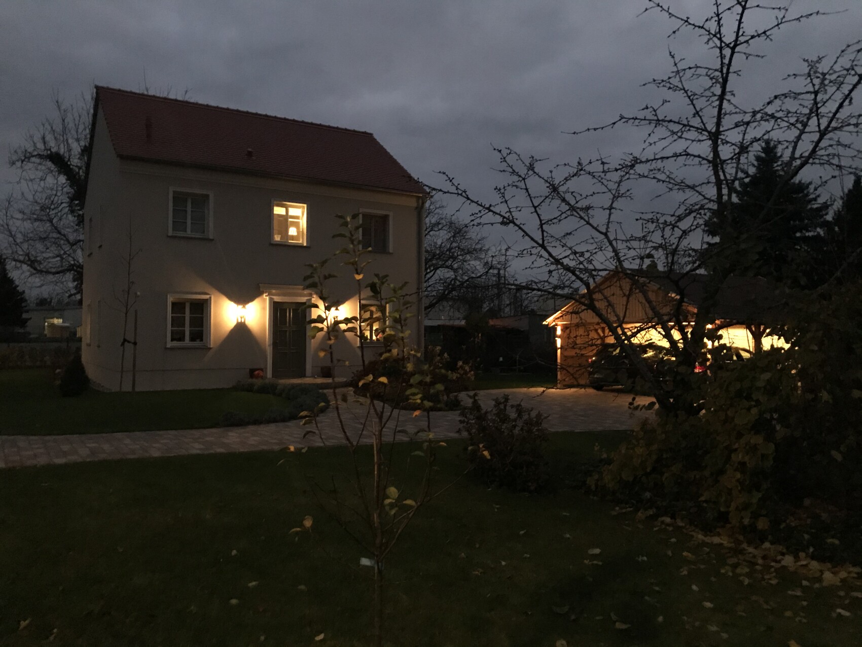 Zweistöckiges Haus bei Dämmerung mit warmem Licht an Eingangstür, Gartenweg und Garage.