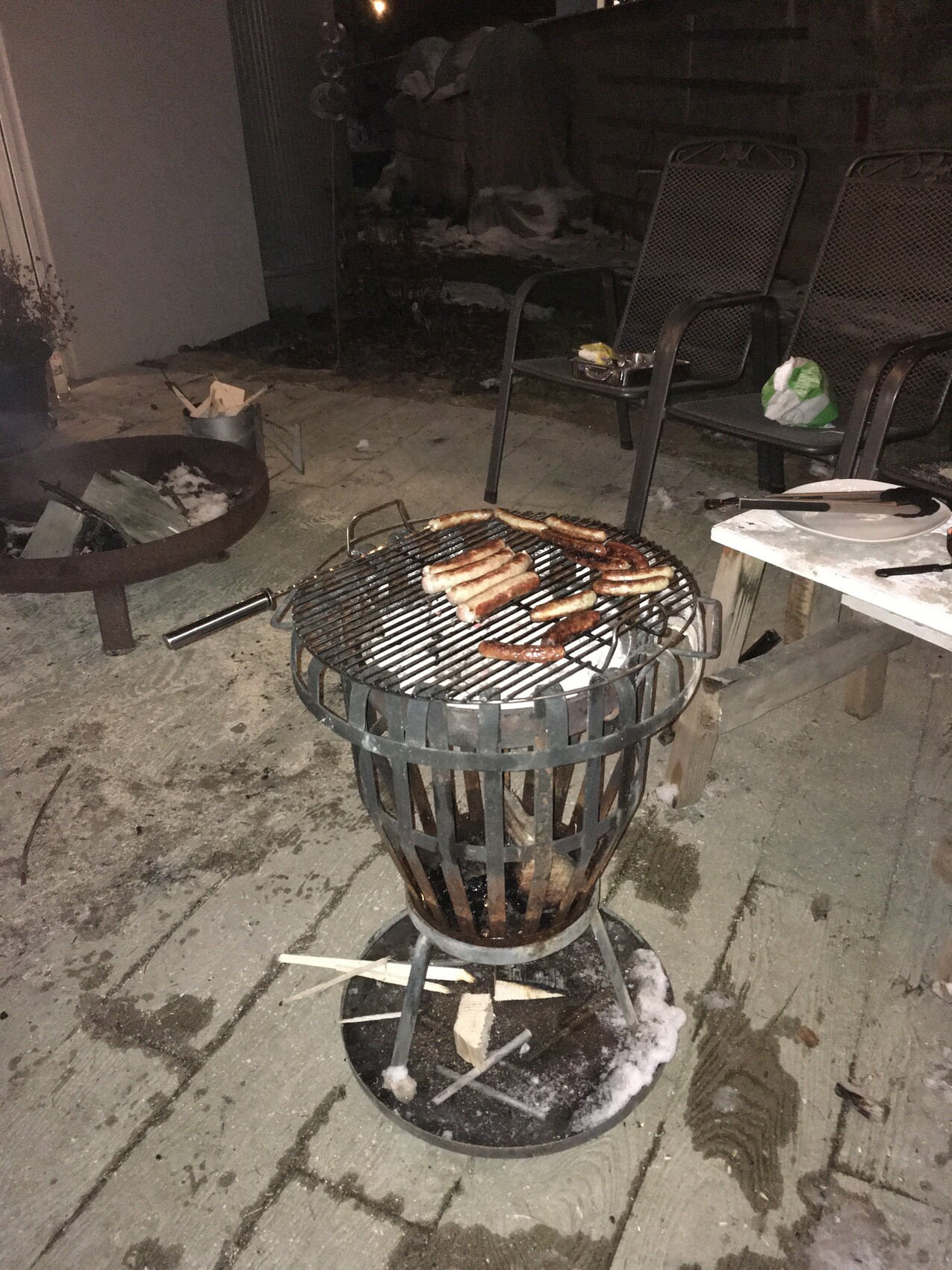 Runder Metallgrill mit Würstchen, daneben Feuerstelle und Gartenstühle bei Nacht