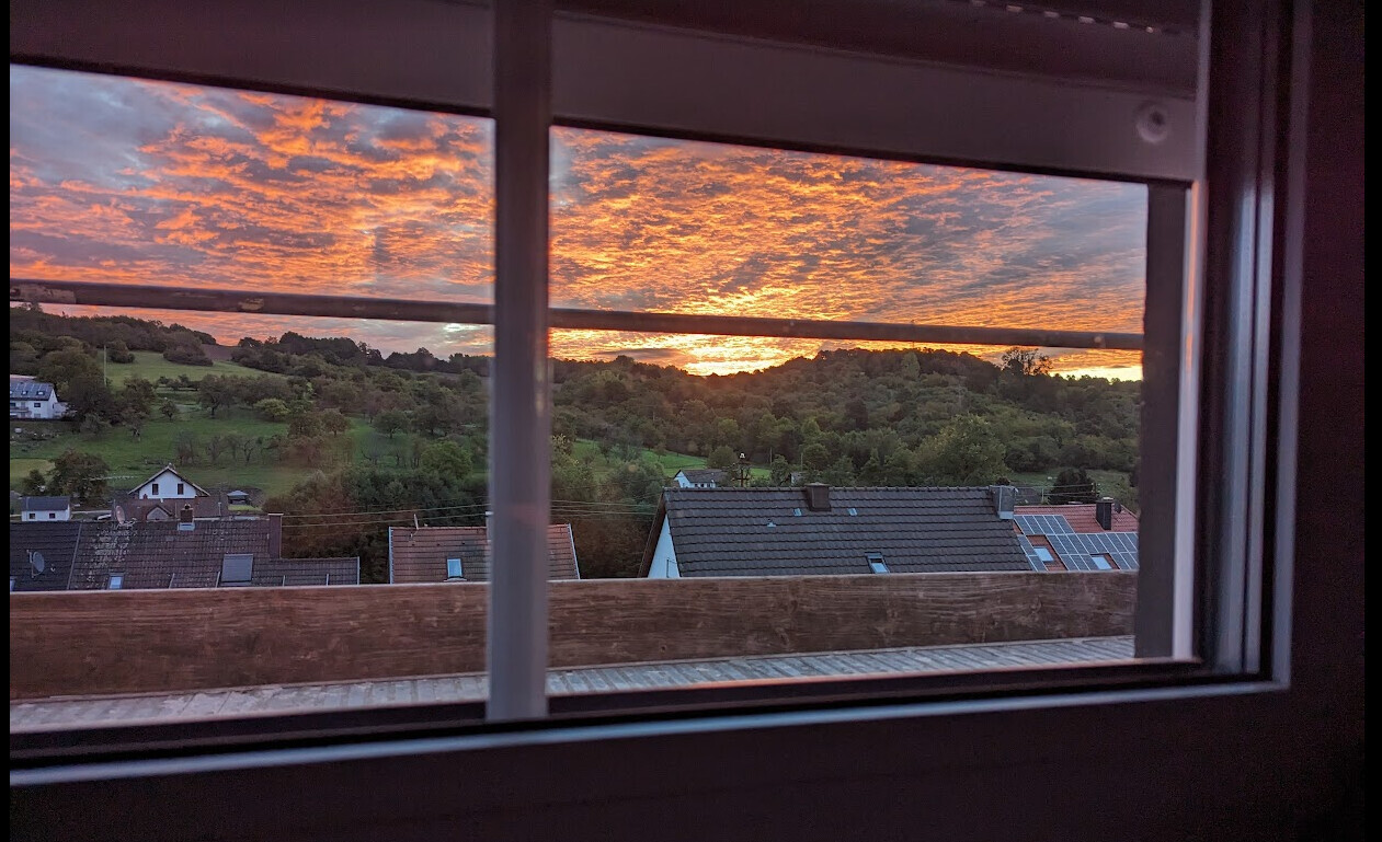 Sonnenuntergang hinter Dächern und grünen Hügeln, durch Fenster gesehen