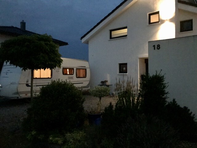 Nächtliche Szene: weißes Haus Nr. 18 mit beleuchtetem Fenster und links Caravan im Garten.