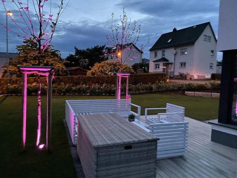 Abendliche Gartenszene mit weißen Korbmöbeln auf Holzdeck, rosa Beleuchtung an Bäumen.