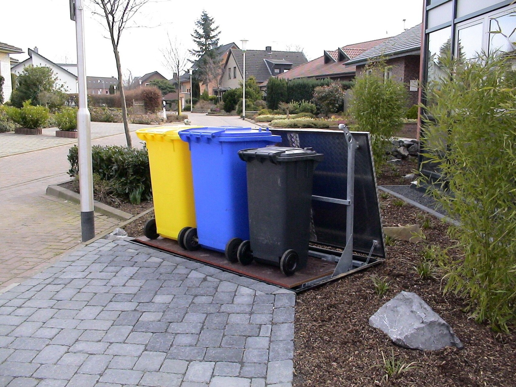 Mehrere Mülltonnen in Gelb, Blau und Schwarz stehen auf einer Metallplattform neben einem Gebäude.