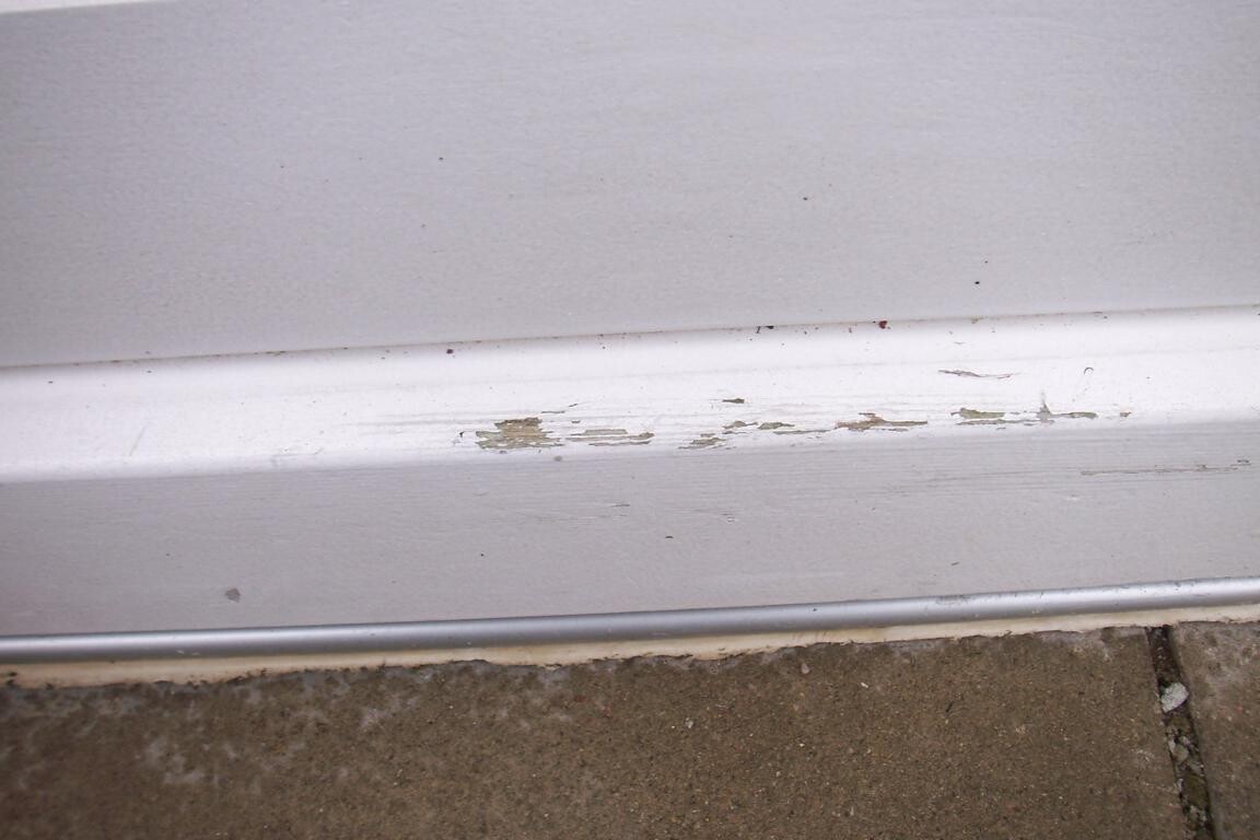 Nahaufnahme einer weißen Türunterkante mit abgeblättertem Lack über Betonboden.